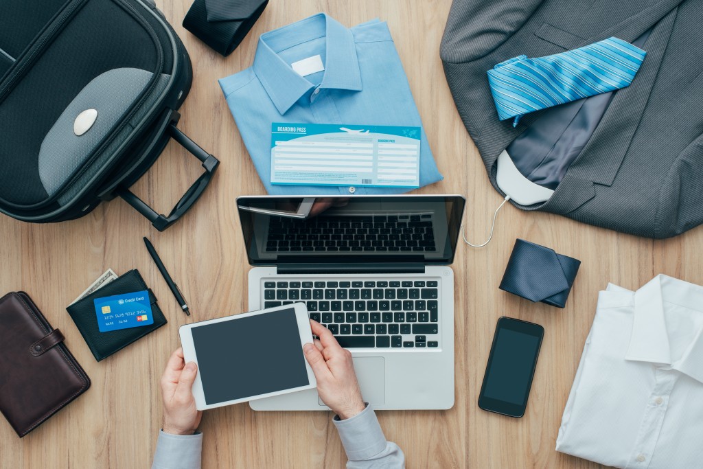 Corporate businessman packing his bag and planning a business trip, he is booking flights online using a digital tablet, travel and technology concept
